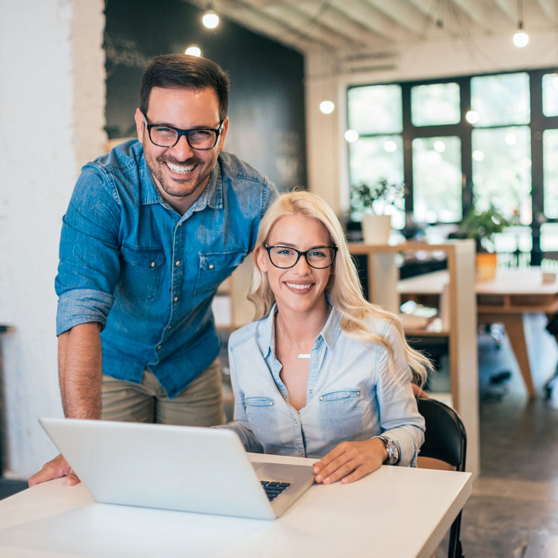 A man (standing) and a woman (sitting) at a desk with a laptop.