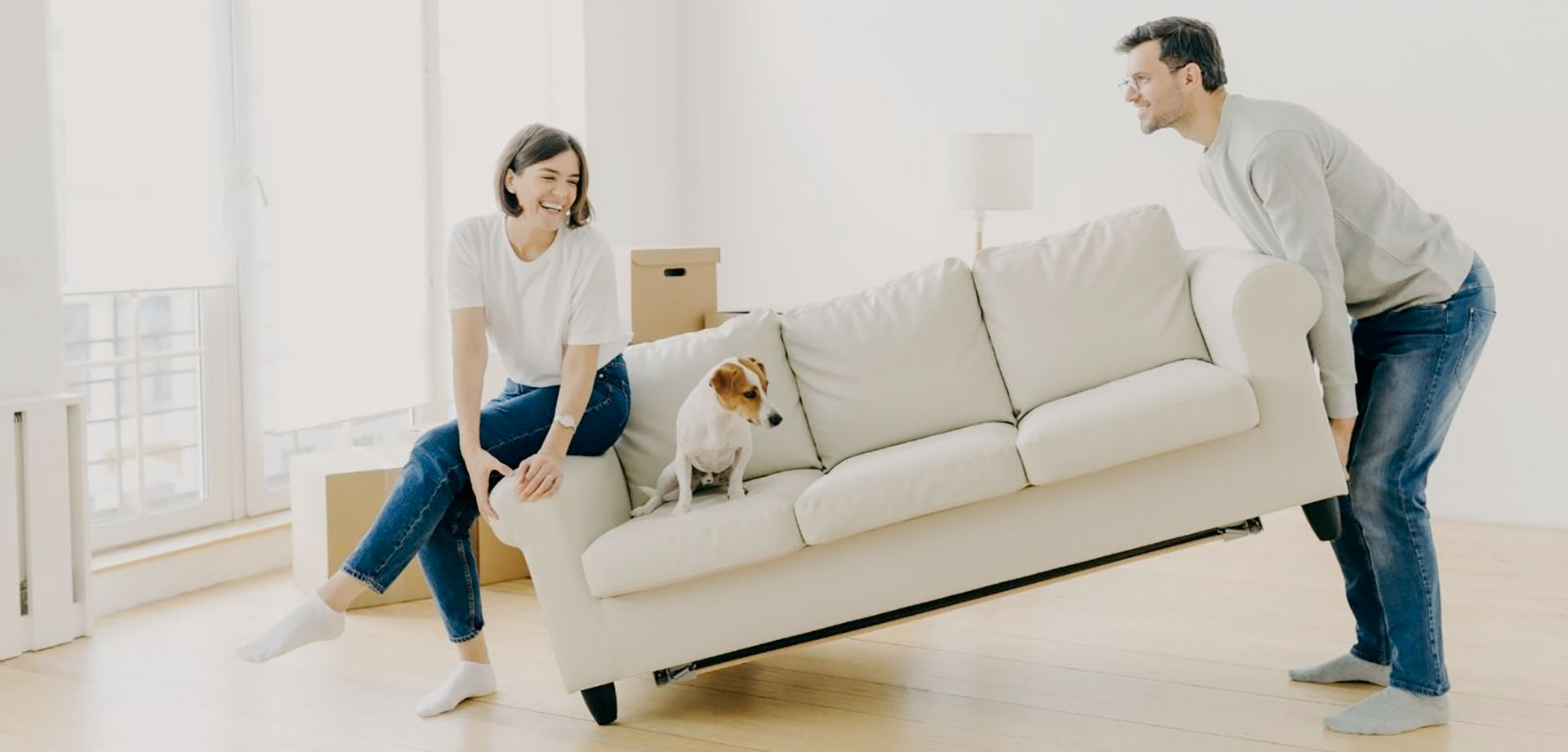 A couple moving a sofa. The man has lifted one end while the female sits on the other end with their dog