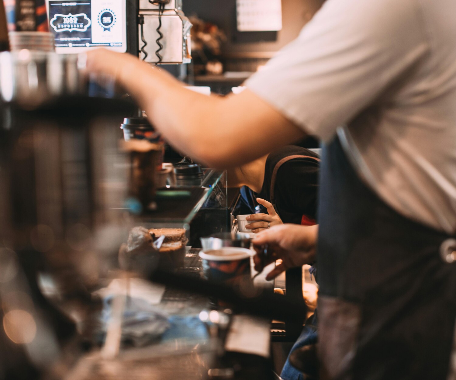 Barista behind a busy café counter preparing drinks, with cups and a pastry in view (motion blur)