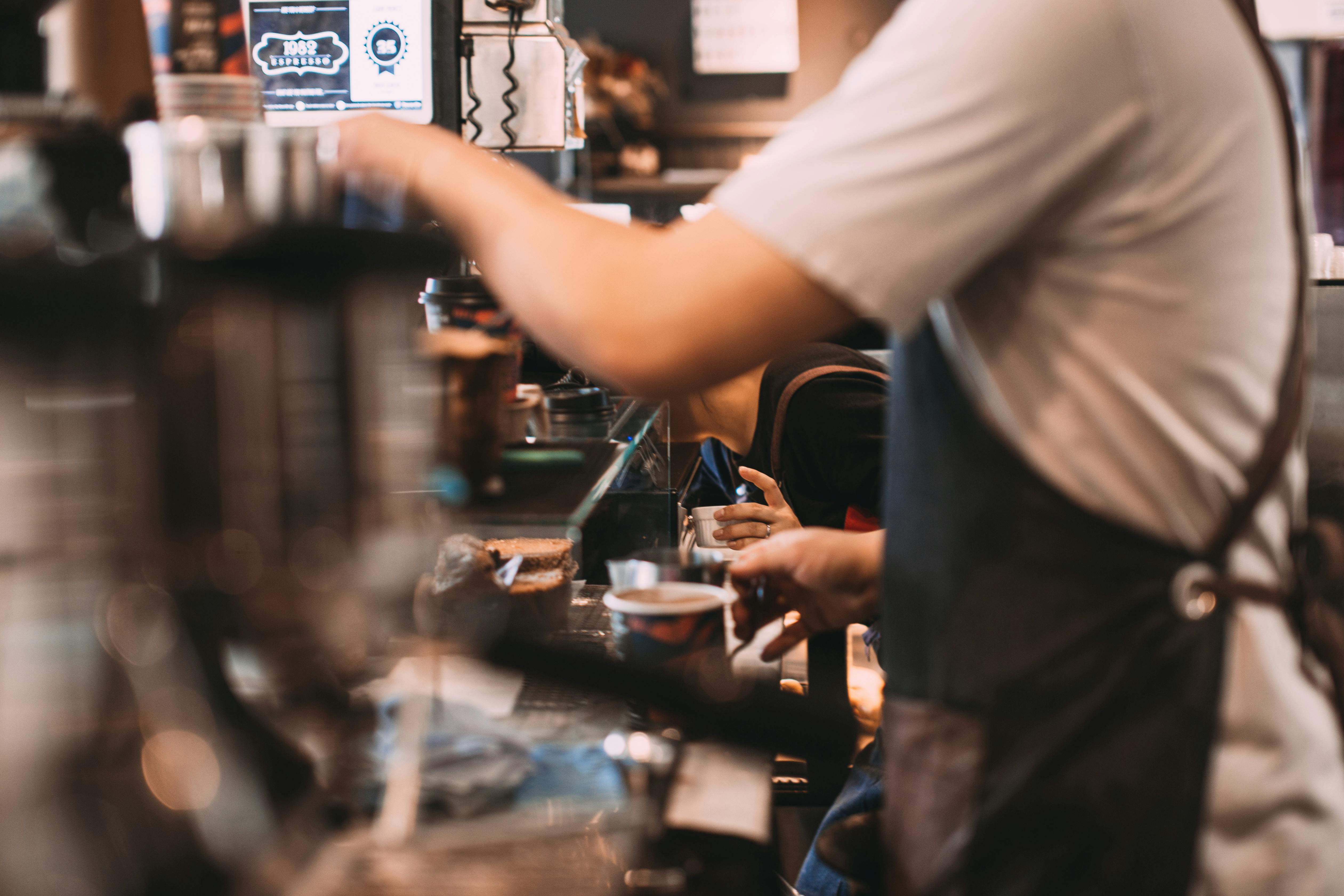 Barista behind a busy café counter preparing drinks, with cups and a pastry in view (motion blur)