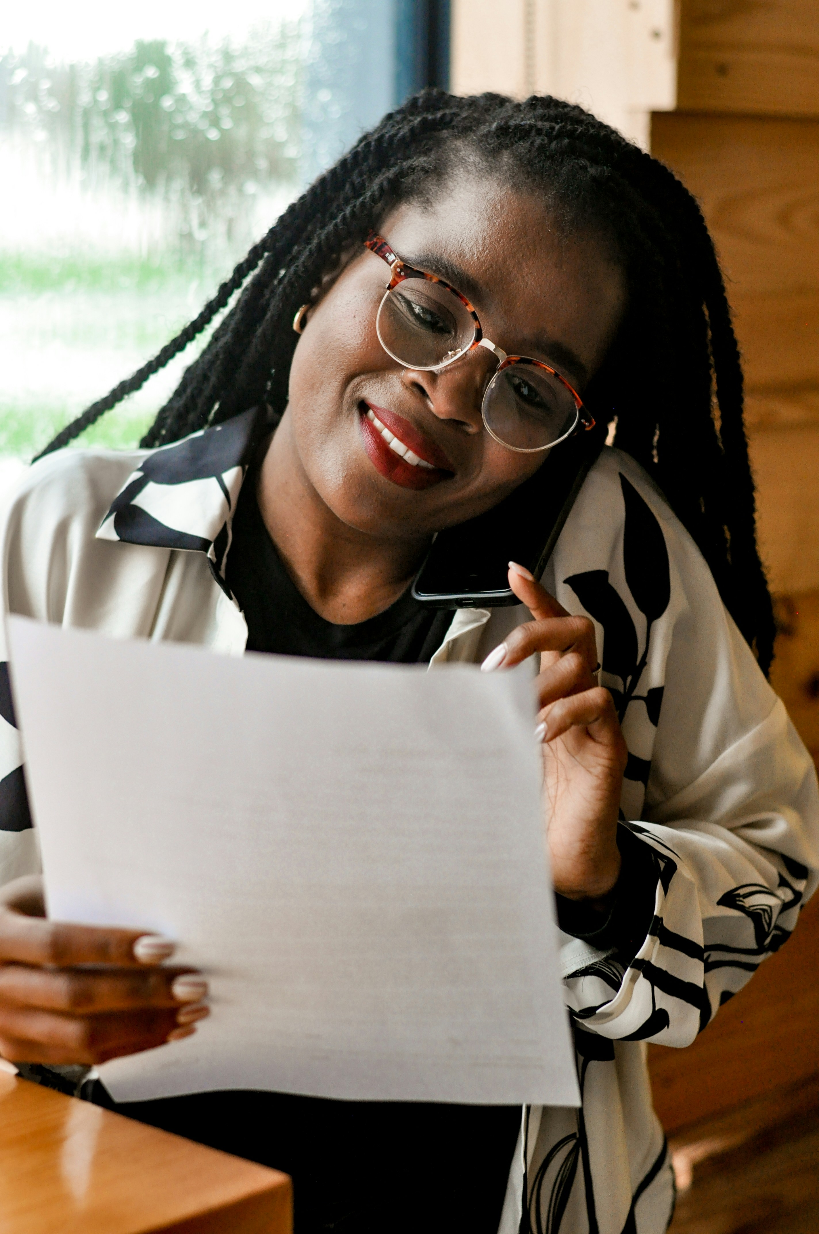 first home buyer phone call - UFINANCIAL Woman with tortoiseshell glasses smiles while holding a sheet of paper and a smartphone to her ear/sweet moment of focused reading at a desk.
