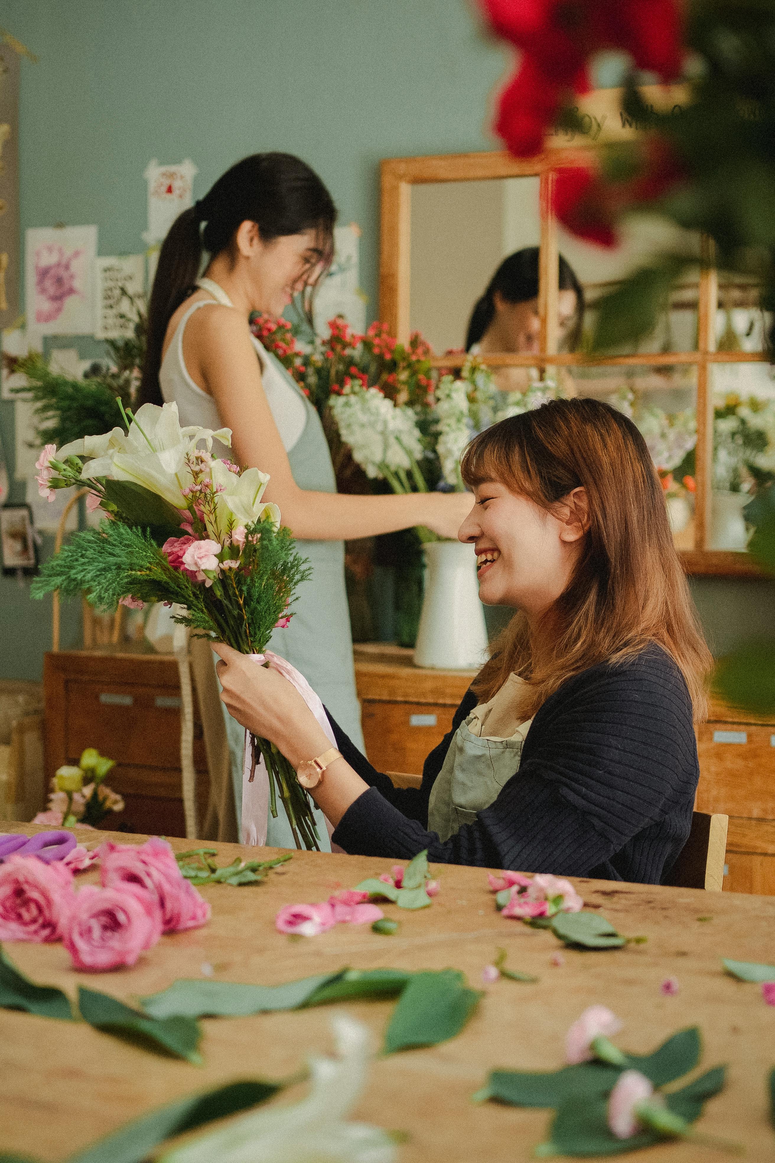 small business florist employee - UFINANCIAL Smiling woman in an apron hands a bouquet of pink and white flowers in a florist shop. Behind her, another florist arranges blooms.