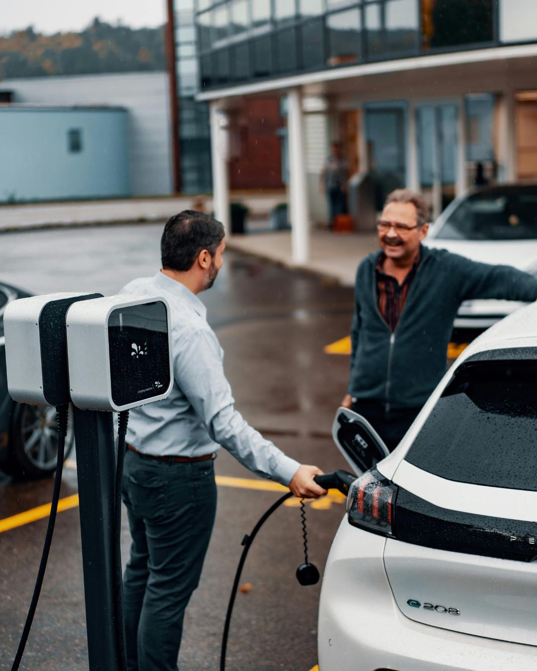 Two men charging a white electric car at a public charging station in a parking lot, rain on the ground.