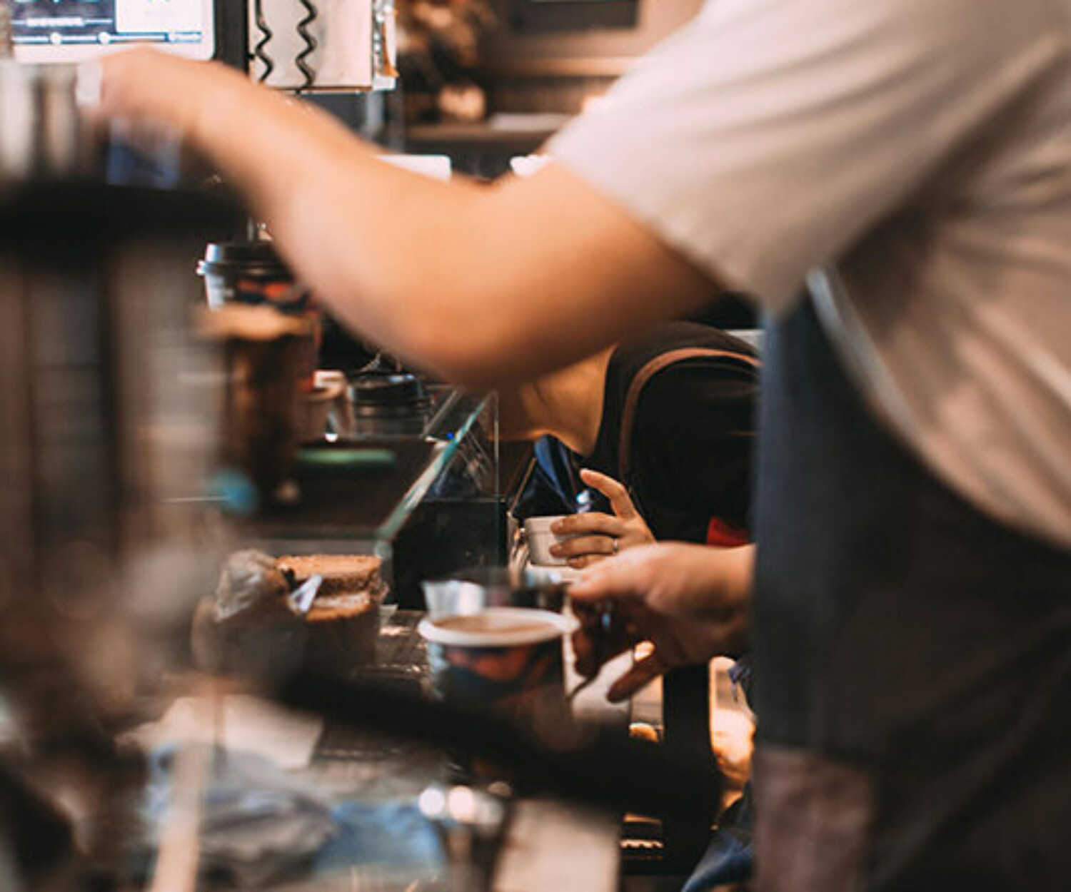 Barista in an apron serves a coffee at a busy cafe counter while customers reach for drinks