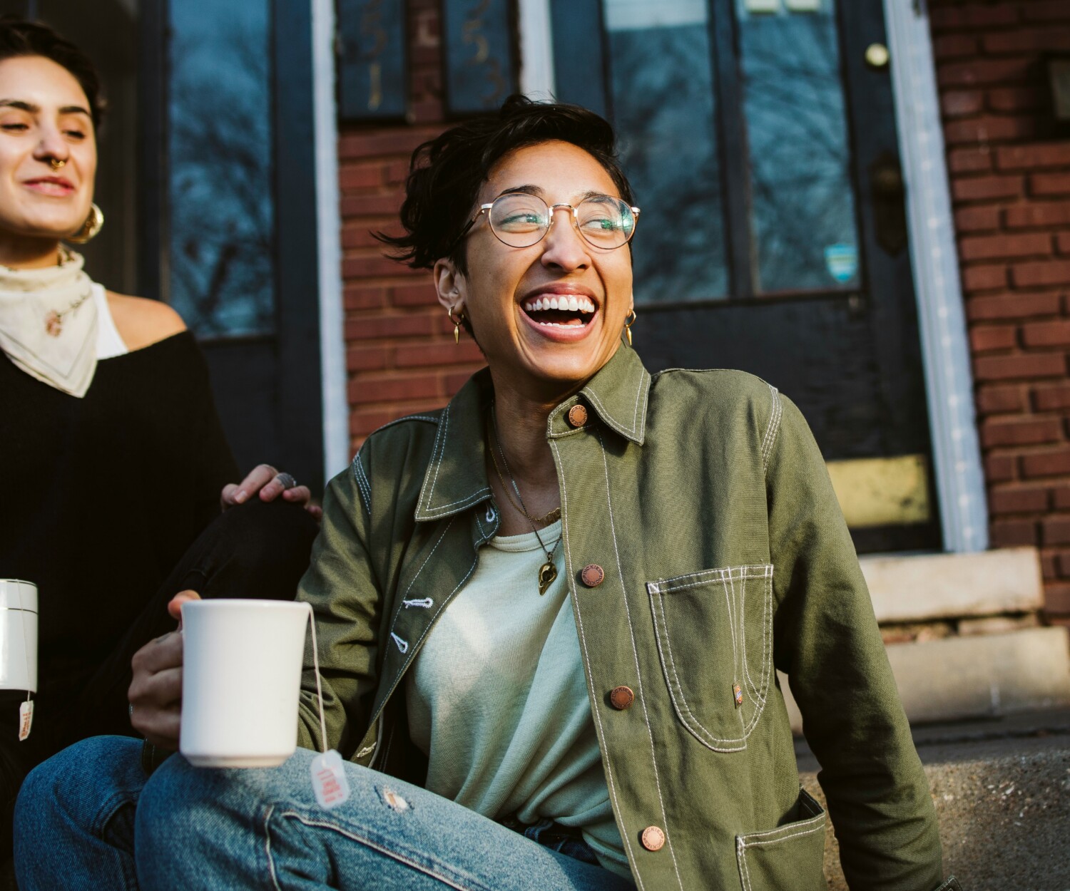 Two friends sit on brick steps outside, laughing and holding white mugs during a casual outdoor moment.