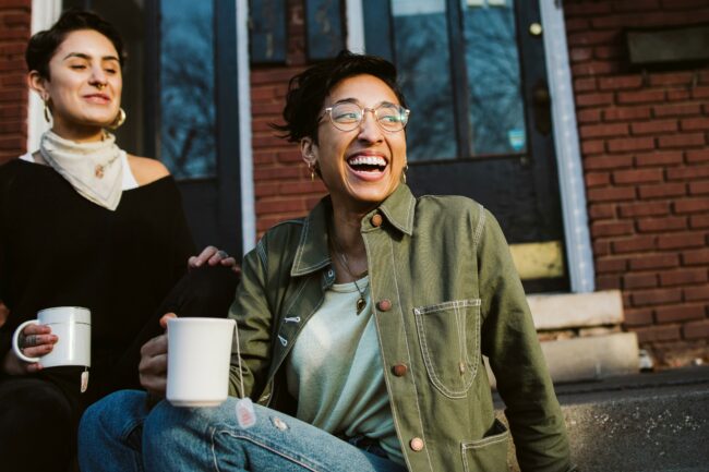 Two friends sit on brick steps outside, laughing and holding white mugs during a casual outdoor moment.