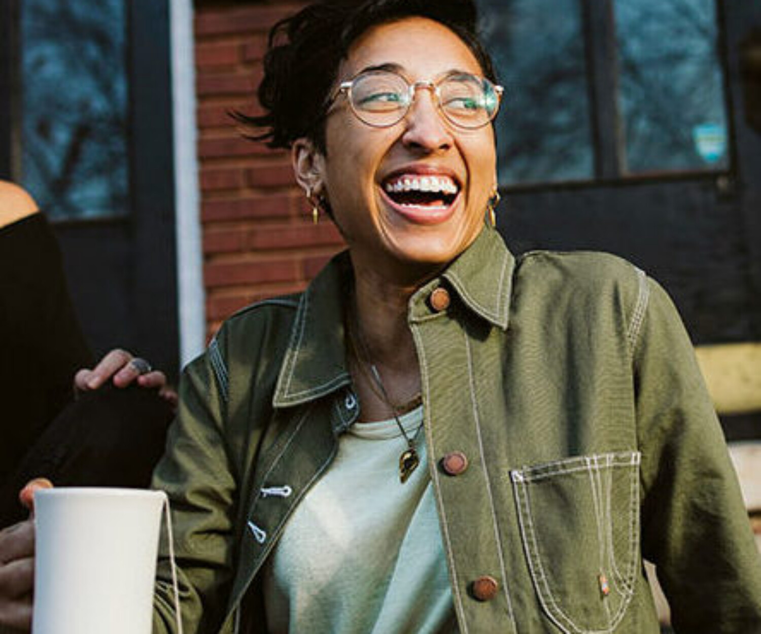Two women sitting on concrete steps in front of a brick building, laughing and holding white mugs, enjoying coffee together.