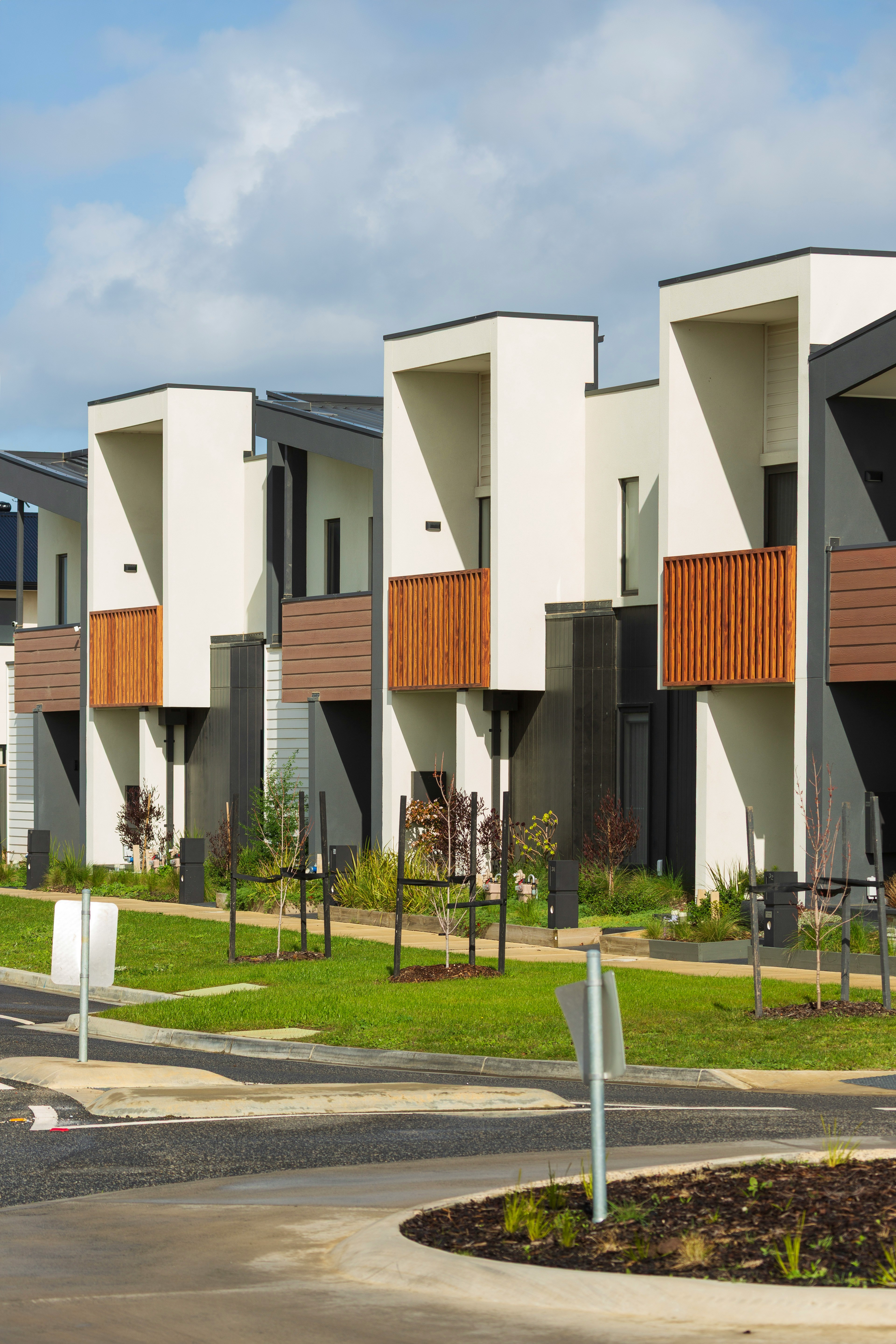 subdivided properties in victoria - UFINANCIAL Row of modern white townhouses with wooden balconies under a blue sky, adjacent to a curved road and landscaped green space