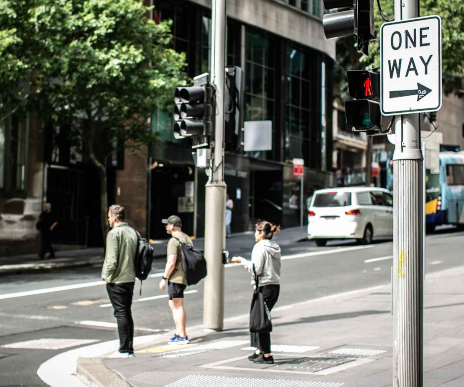 Pedestrians wait to cross at a city intersection beside a 'One Way' sign and traffic lights, with a bus in the background.