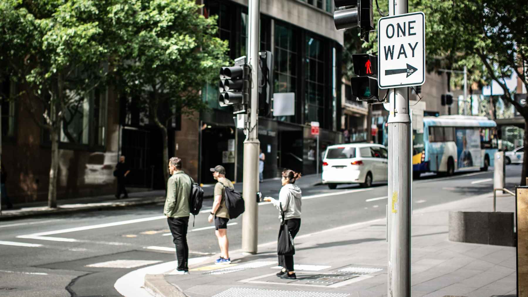 Pedestrians wait to cross at a city intersection beside a 'One Way' sign and traffic lights, with a bus in the background.