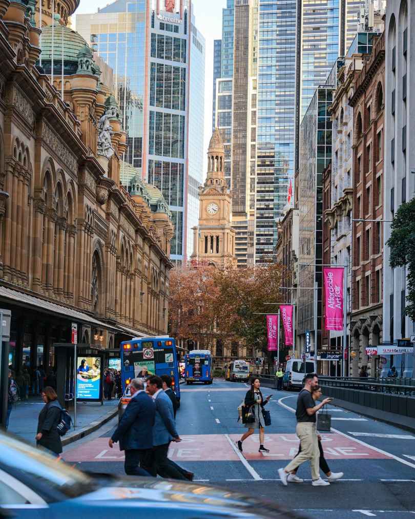 Busy city street with pedestrians crossing in front of a mix of historic sandstone buildings and modern skyscrapers; a clock tower in the distance.