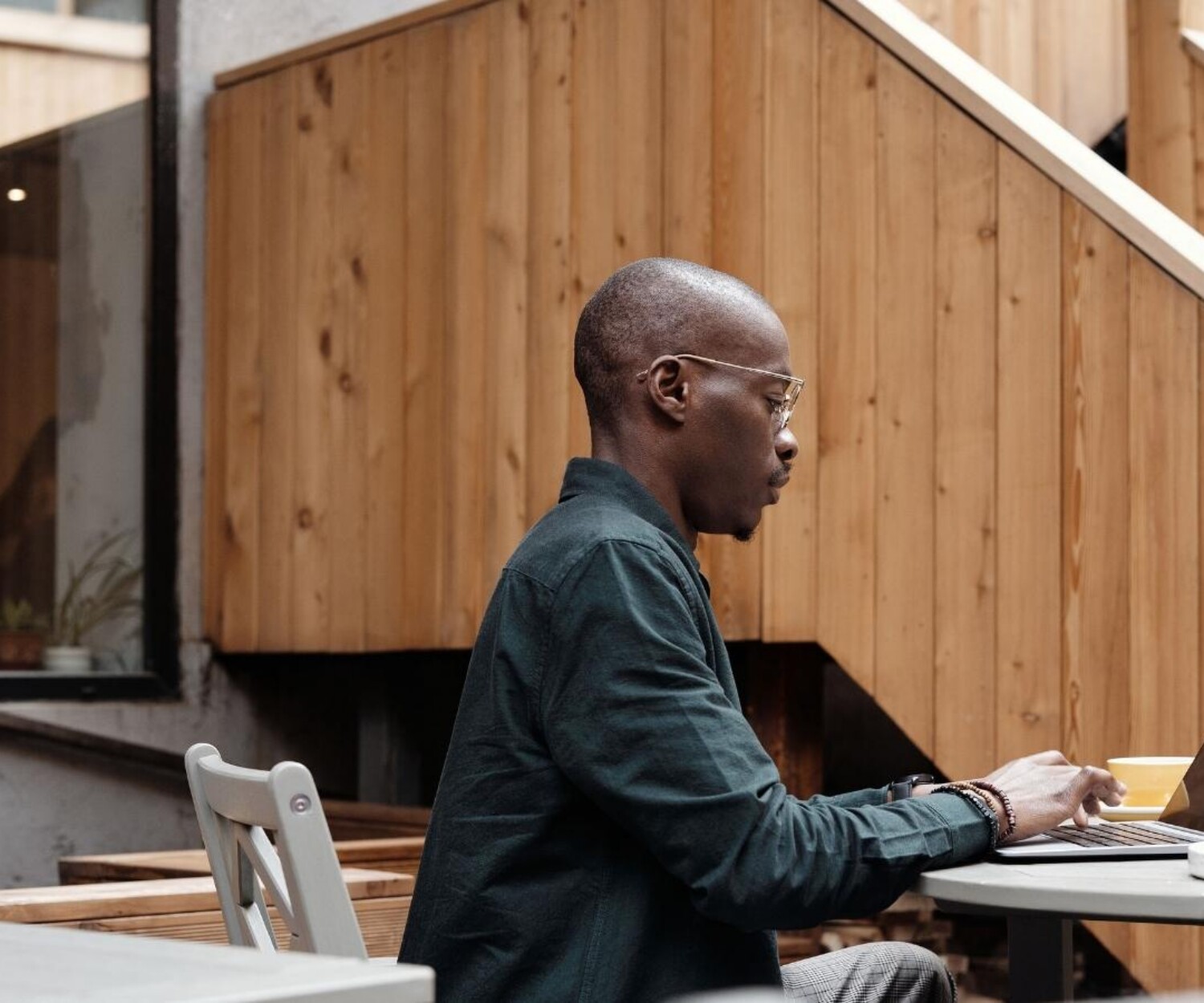 Man wearing glasses sits at an outdoor cafe table, typing on a laptop with a pastry on a plate nearby and a wood-paneled wall in the background.