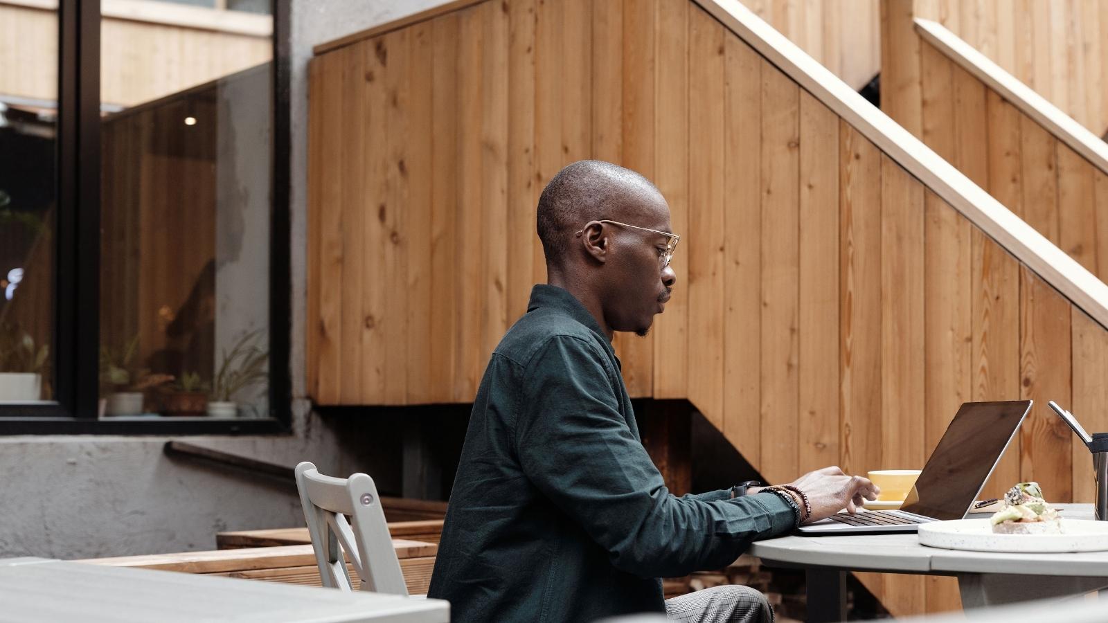 Man wearing glasses sits at an outdoor cafe table, typing on a laptop with a pastry on a plate nearby and a wood-paneled wall in the background.