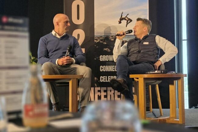 Chris Judd and Kris Court sit on a stage in a panel discussion, each holding a microphone, with a large UFfinancial banner behind them and a blurred conference table in the foreground.