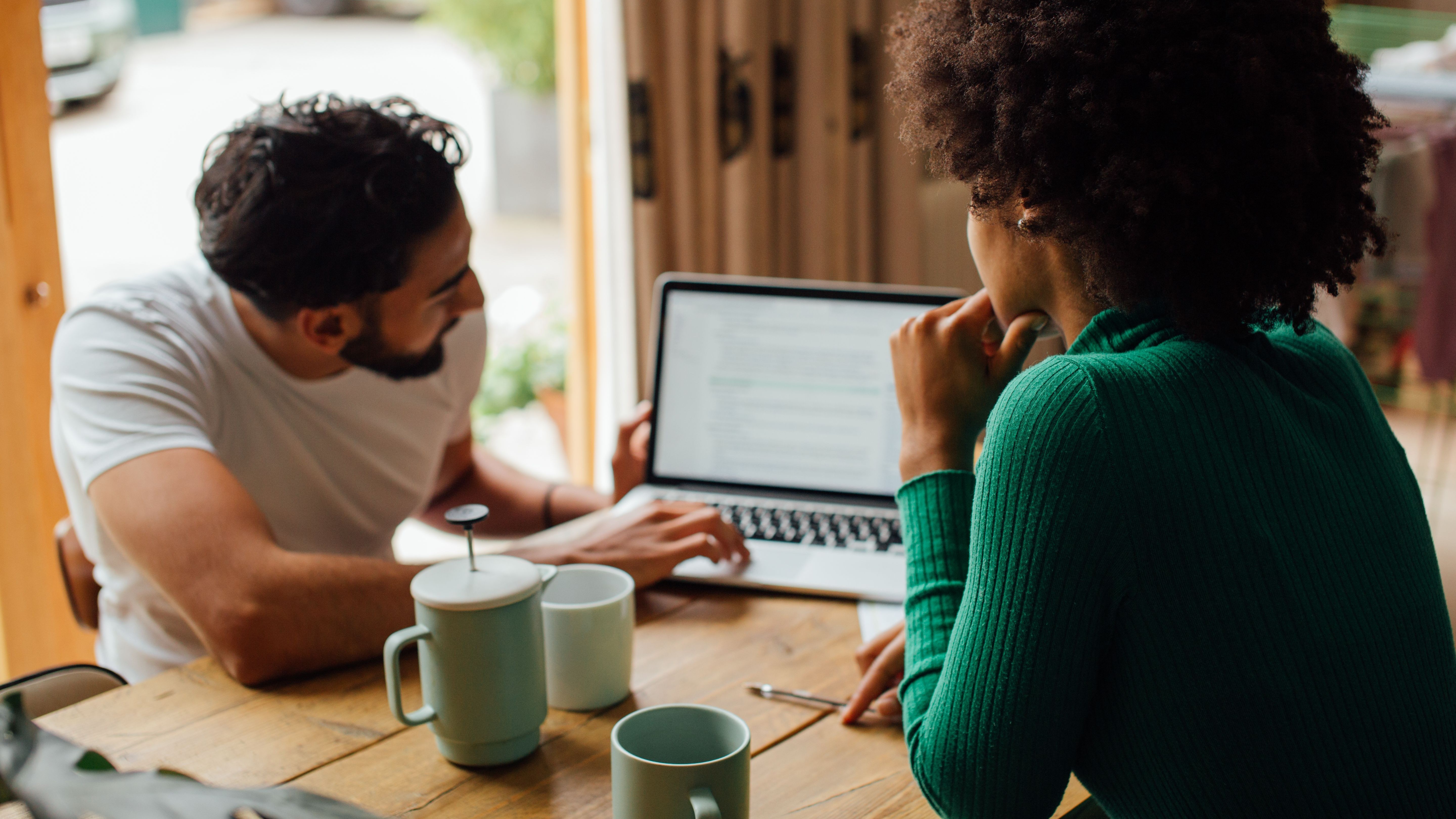 Two people sit at a wooden table and look at a laptop screen, with coffee mugs nearby.