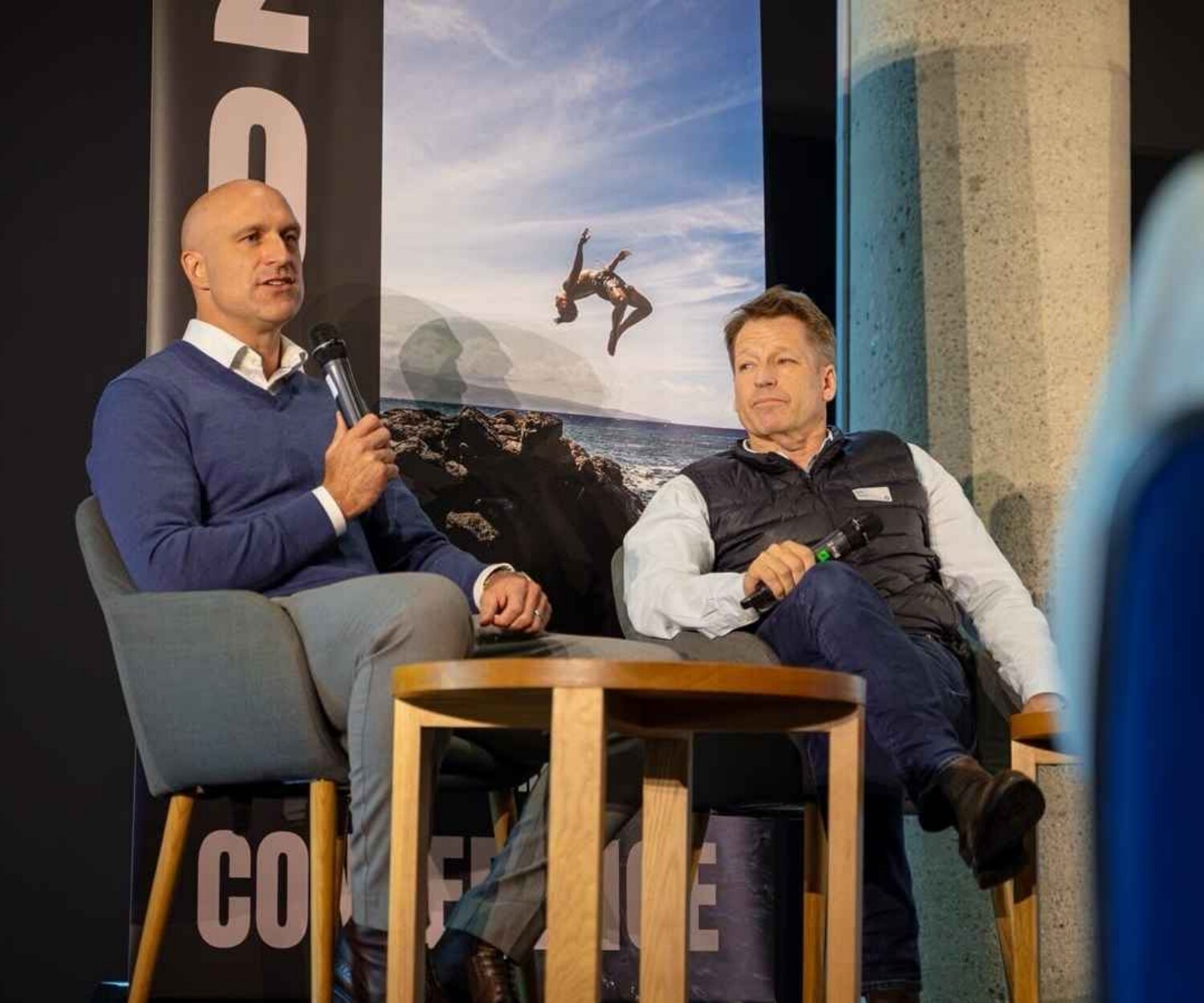 Two men sit on stage in a panel discussion, one speaking into a microphone while the other listens.