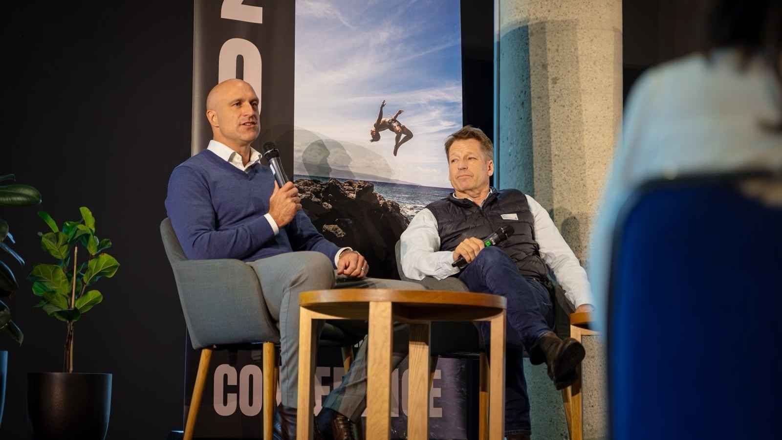 Two men sit on stage in a panel discussion, one speaking into a microphone while the other listens.