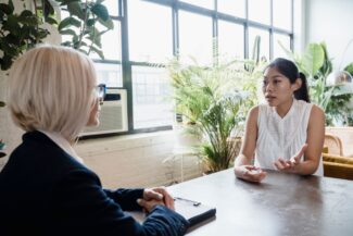Two colleagues discuss at a table in a bright, plant-filled office; one woman gestures while the other sits with her back to the camera.