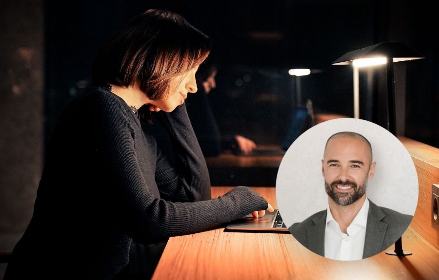 Woman typing on a laptop at a wooden desk in dim light; a circular portrait of a smiling man in a suit sits nearby.