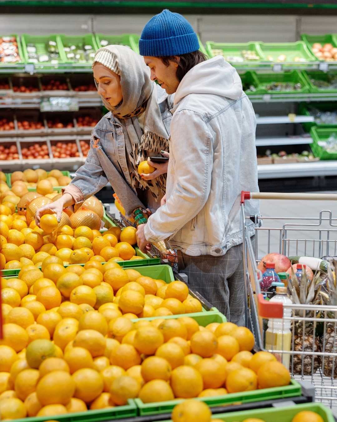 Two shoppers in a grocery store select oranges from bright green crates in the produce section, leaning over the display to pick fruit.