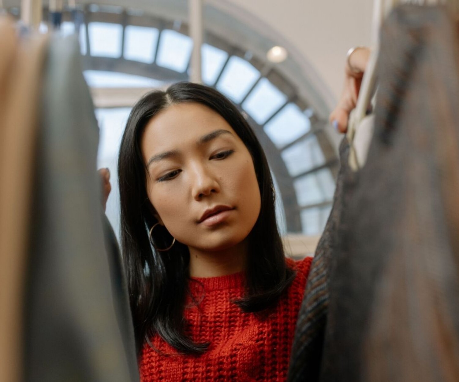 Woman in a red knit sweater browses hanging coats in a clothing store, inspecting garments closely.