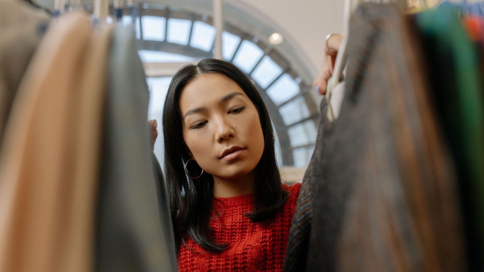 Is buy now pay later a good idea header image - UFINANCIAL Woman in a red knit sweater browses hanging coats in a clothing store, inspecting garments closely.