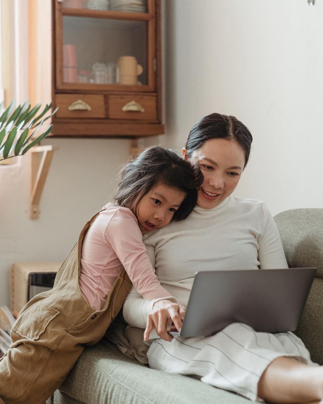 A girl hugs her mother on the couch as they look at a laptop.