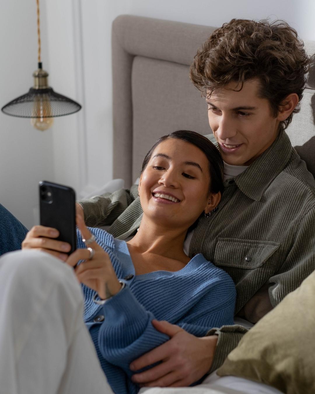 A woman lays on a man's chest and shows him something on her phone.