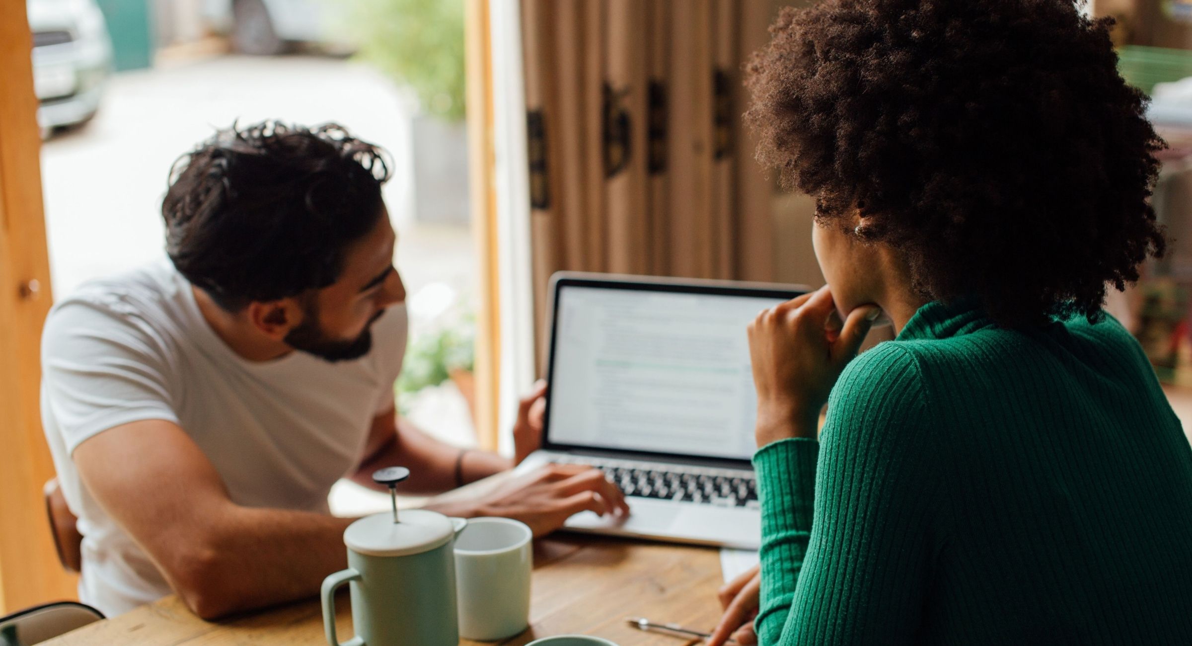 Two people sit at a wooden table and look at a laptop screen, with coffee mugs nearby.