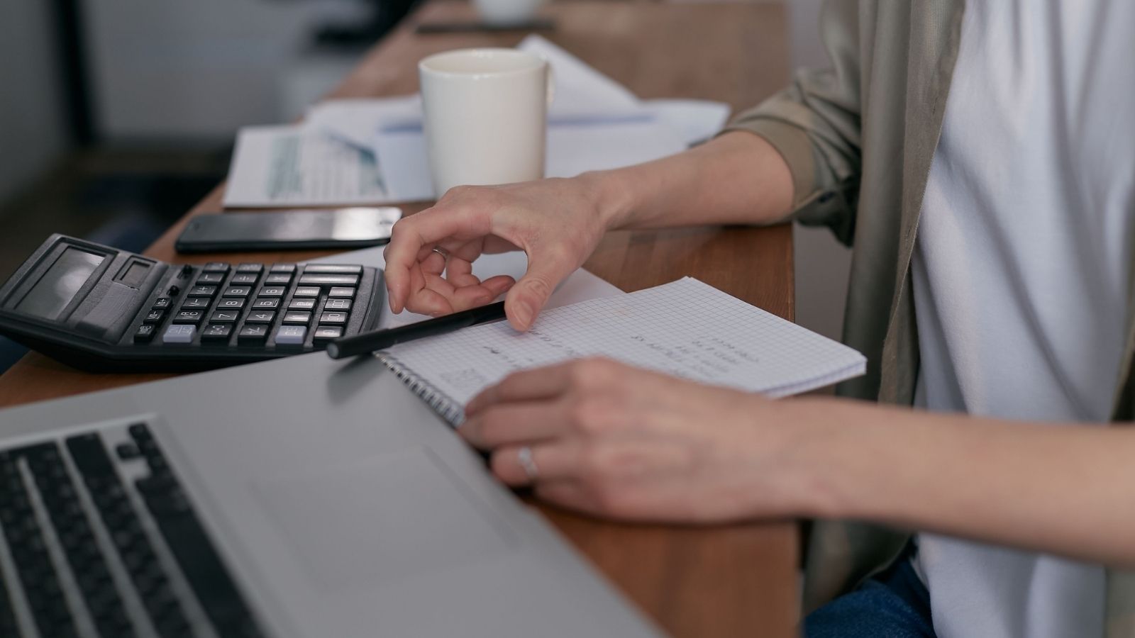 Person sits at a desk, using a calculator while writing in a notebook; a laptop, phone, and coffee mug are nearby.