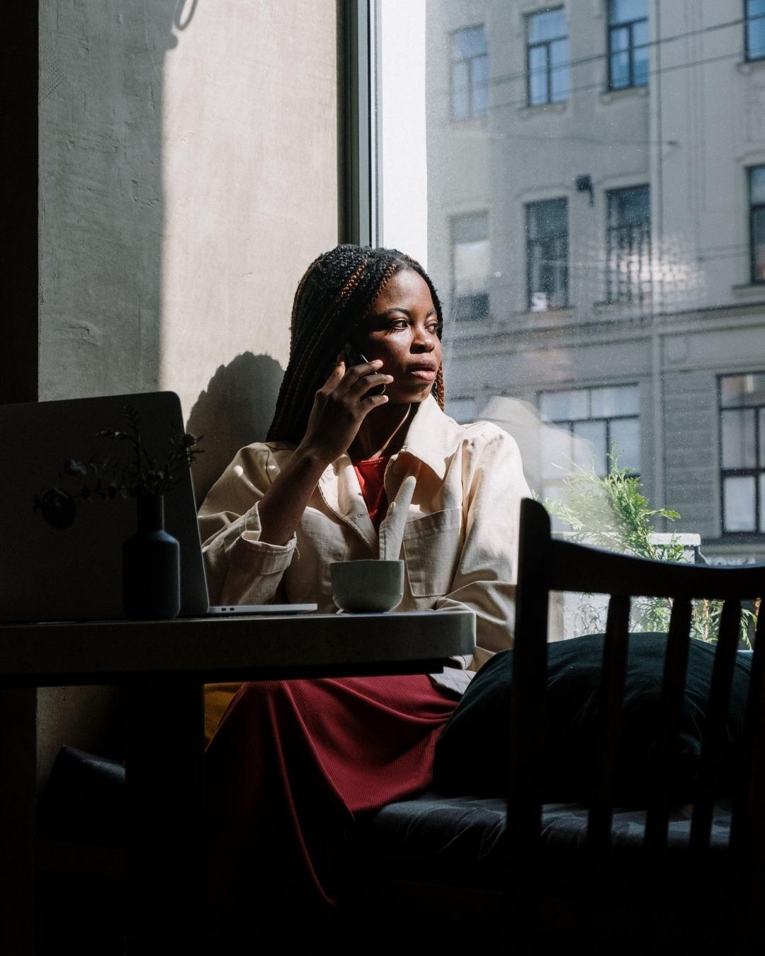 Woman in a beige jacket and red skirt sits at a cafe table by a window, talking on her cellphone with a laptop and cup on the table.