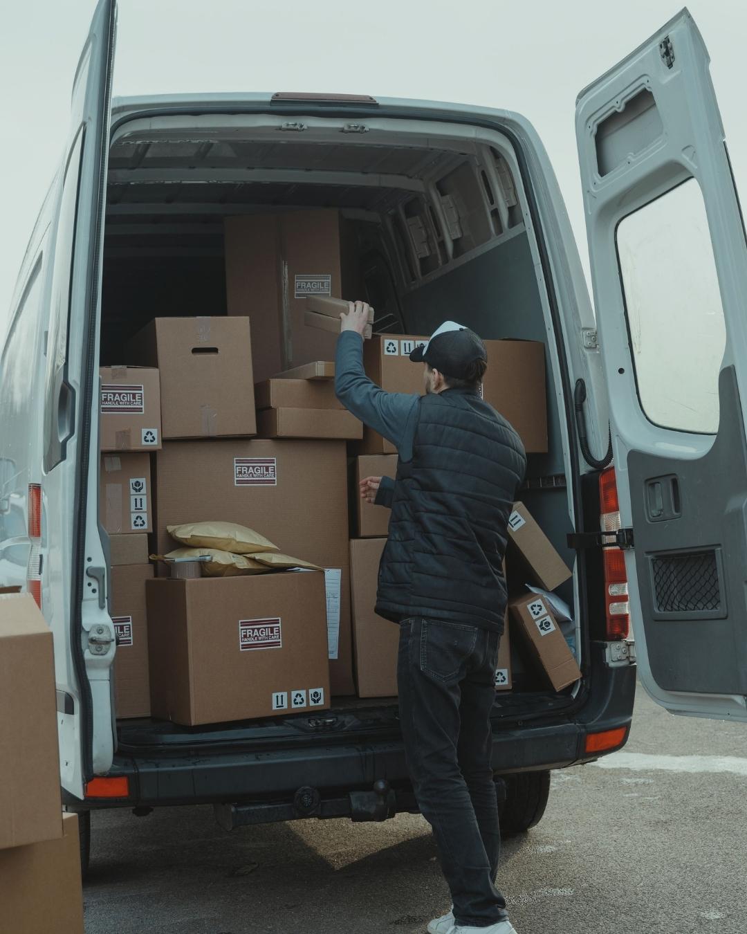 A person loading stacked cardboard boxes into the back of a white van, some labeled fragile.